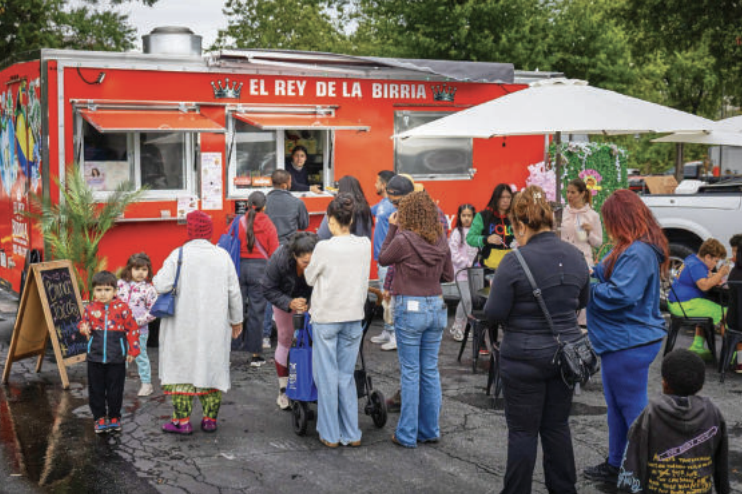 People stand in line for a red food truck titled El Rey De La Birria. A white umbrella stands in front of it.